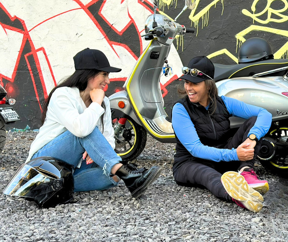two young ladies sitting on the ground, chatting next to their Vespa scooters