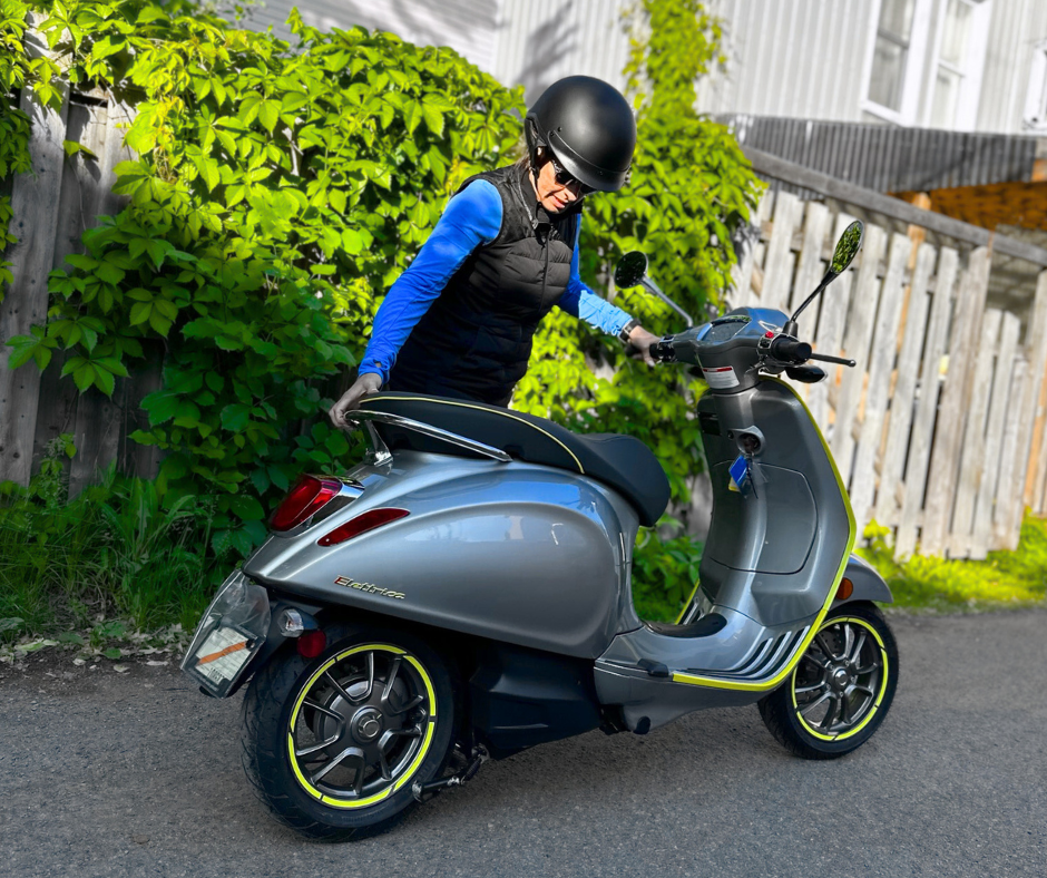 woman standing next to a Vespa in front of a wooden fence covered with greenery